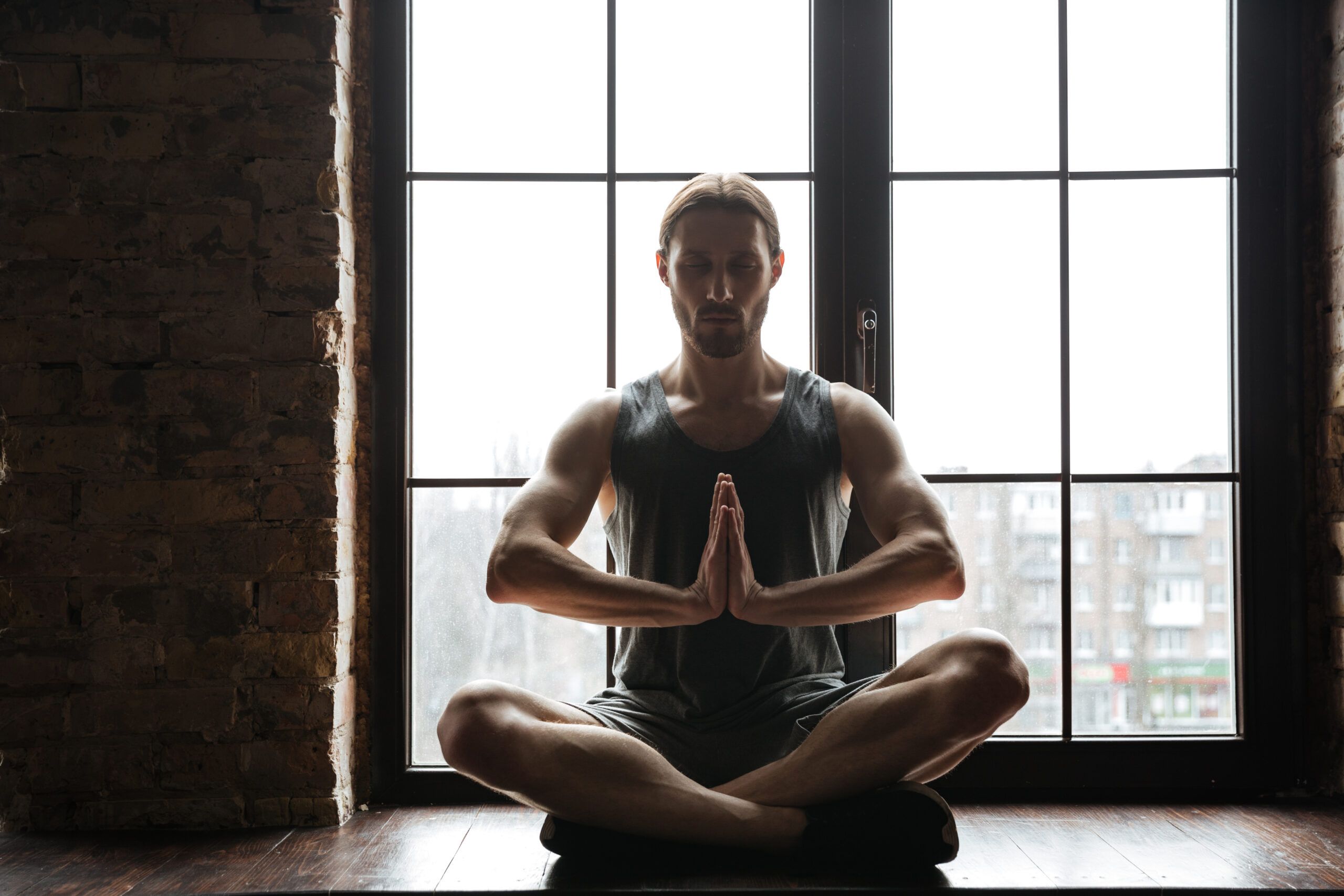 Portrait of a healthy young sportsman meditating in lotus pose with eyes closed while sitting on the windowsill at the gym